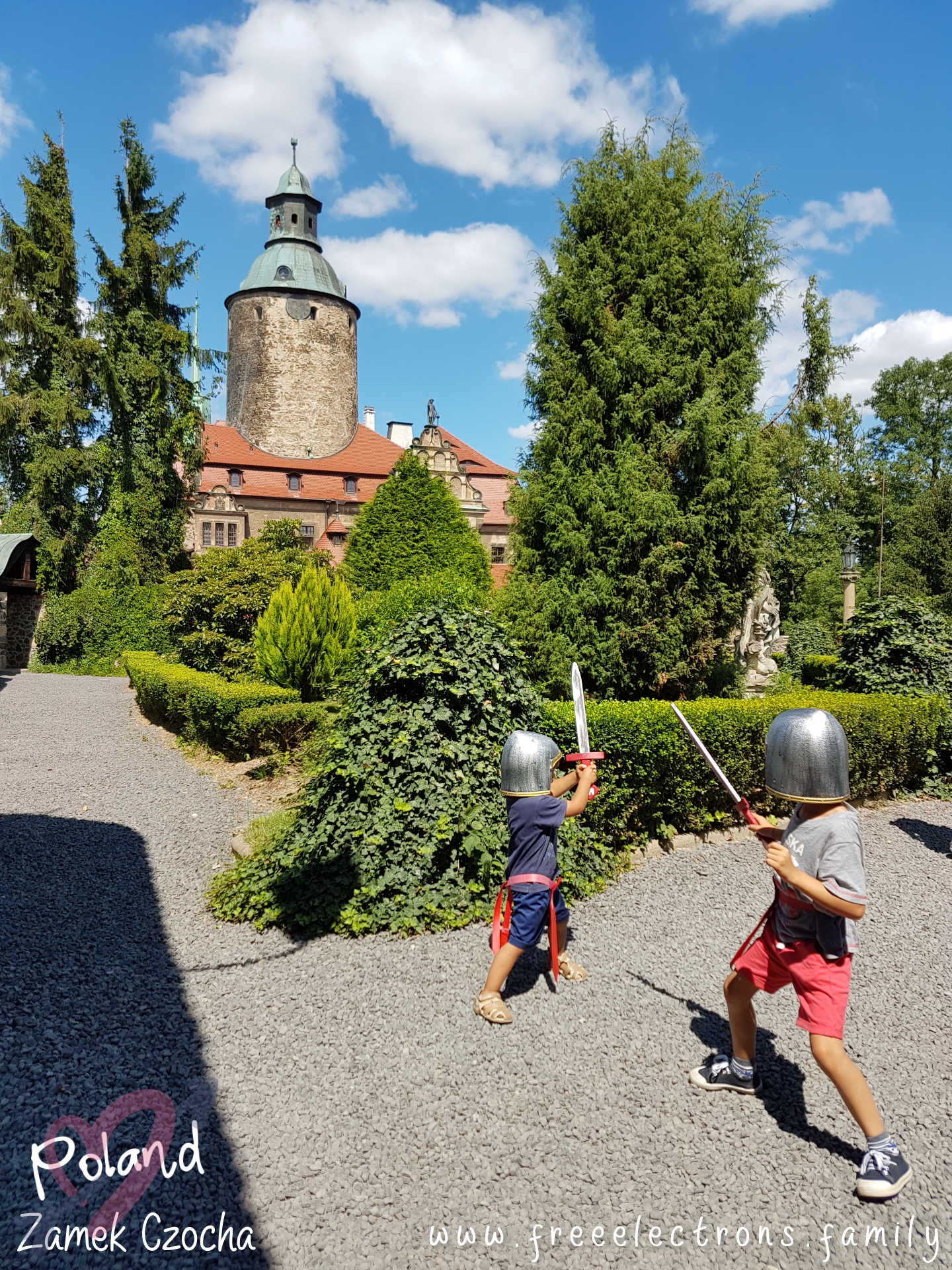 #FreeElectrons.Family - camping road trip Europe Poland, kids at play.

Two young boys in medieval soldier toy helmets and swords playing on castle grounds with lush gardens, under patch-cloudy blue skies.

Text reads: Zamek Czocha Poland (with 2 opaque interlocking hearts behind the text) and www.freeelectrons.com.