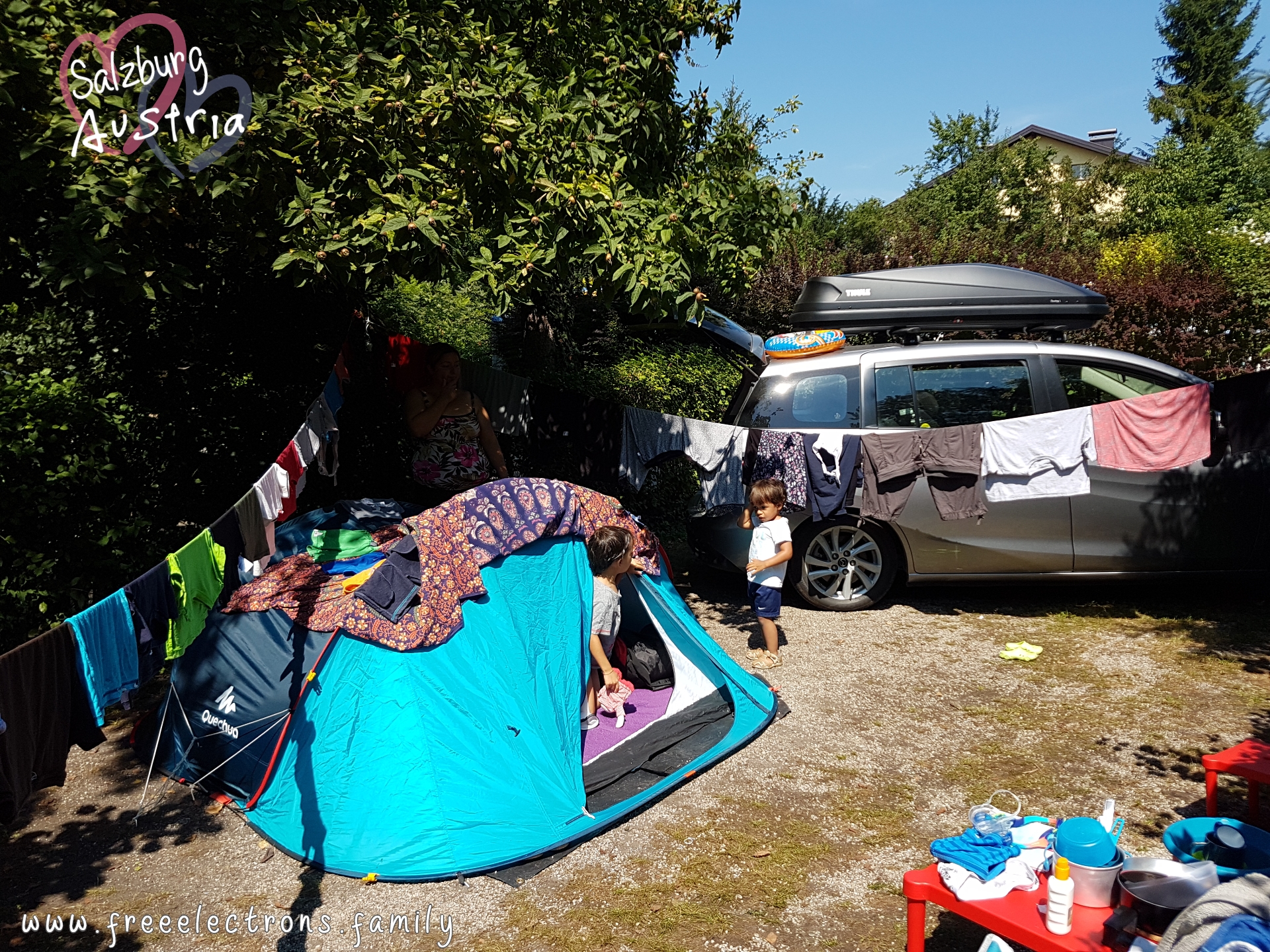 #FreeElectrons.Family - camping road trip Europe, Quechua tent, Austria.

A young boy at the entrance to a light blue Quechua tent talking to a younger boy standing outside.  They are in a small pitch lot surrounded by trees and bushes and flanked by a compact car with a black THULE roof box/luggage on top.  Clothes are drying on clothesline.

Text reads: Salzburg Austria (with 2 opaque interlocking hearts behind the text) and www.freeelectrons.com.