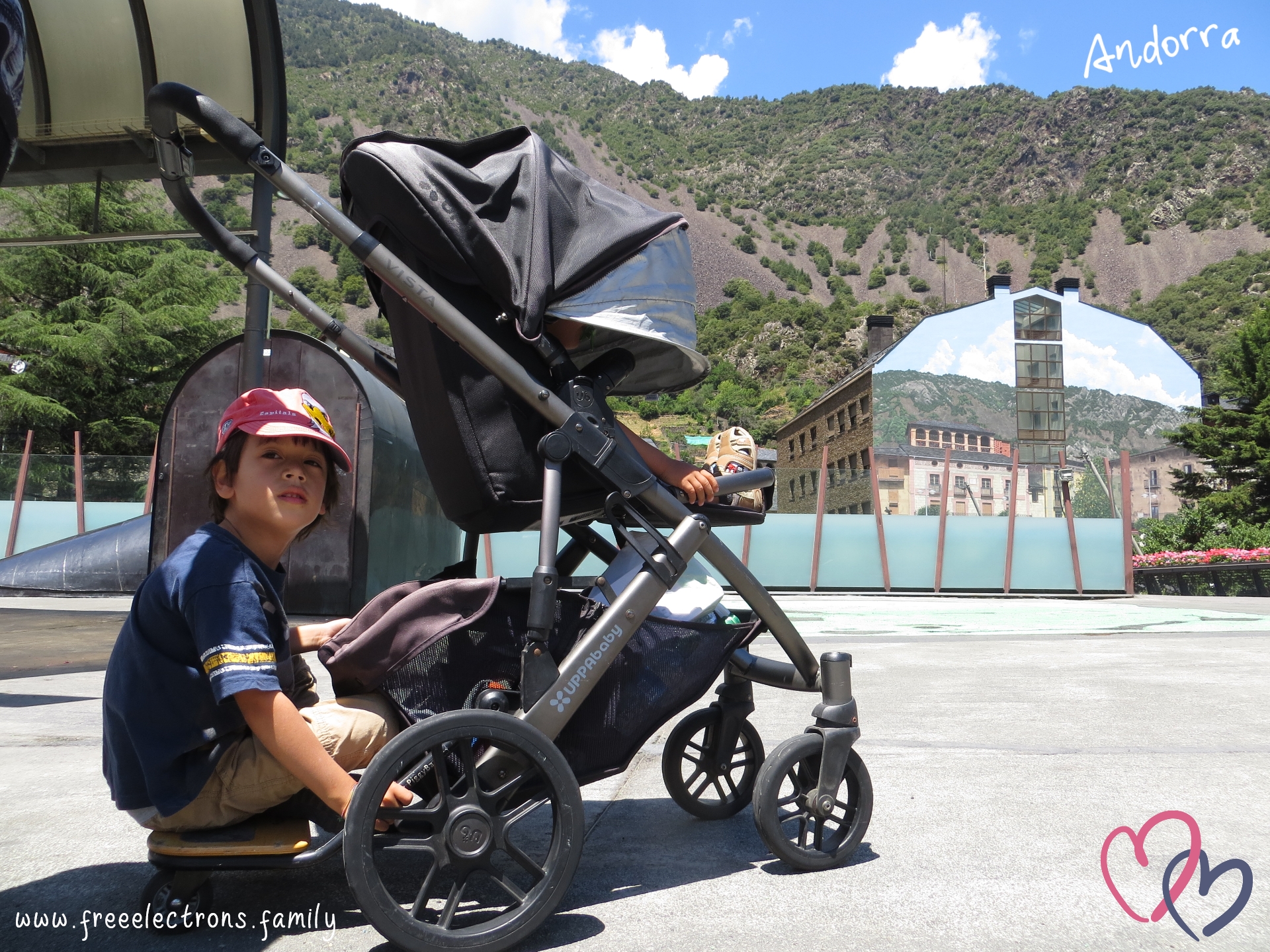 #FreeElectrons.Family - camping road trip Europe, UPPAbaby, kids chilling in Andorra.

Two young kids resting on an UPPAbaby VISTA stroller/pram at an empty plaza.  The older one 
is sitting on the piggyback ride along board, looking past the camera.  The other one in the rumble seat.  A building with a landscape painting that blends in the background of green-patched mountain and, above, blue skies with white patches of clouds.

Text reads: Andorra; www.freeelectrons.com