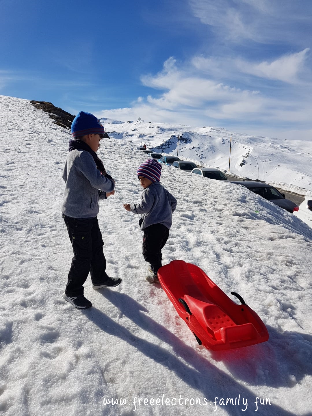 Two young boys with snow caps dragging a red sled up the snow-covered slopes of Hoya de la Mora in the Sierra Nevada, province of Granada, Andalucia, Spain. Andalusian blue sky in the background with swirly clouds

Caption reads: Onwards! Upwards!  To the peak...  Pico de Veleta  (well, maybe not this time).

label: www.freeelectrons.family fun