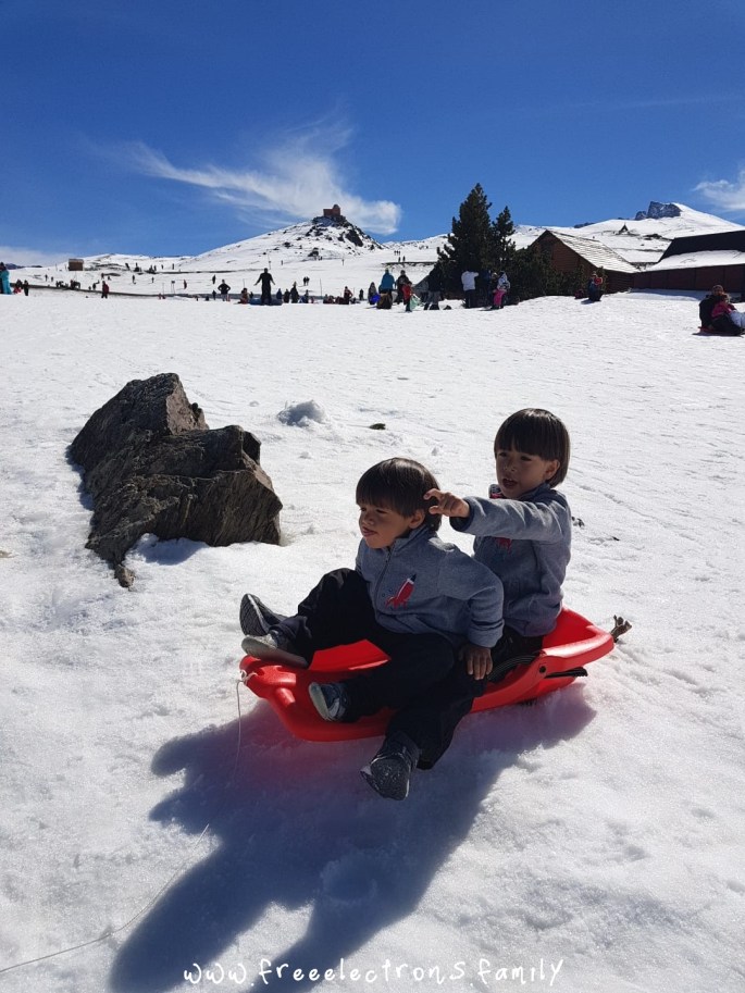 Two young boys on a red sled coming down a slope. One is pointing outward. The other one upfront looks directly and bravely ahead, holding onto to his older brother. In the background. blue sky over the peaks of the mountain atop Hoya de la Mora, in Sierra Nevada, province of Granada in Andalucia, Spain.