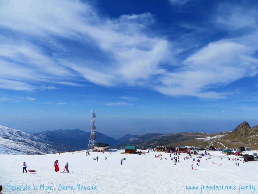 Atop the snow-covered slopes of Hoya de la Mora in the Sierra Nevada, province of Granada, Andalucia, Spain. Children on sleds and various snow plays in the distance. Hazy mountains and plains in the background, under an Andalusian blue sky with swirly clouds. Label: Hoya de la Mora, Sierra Nevada; www.freeelectrons.family