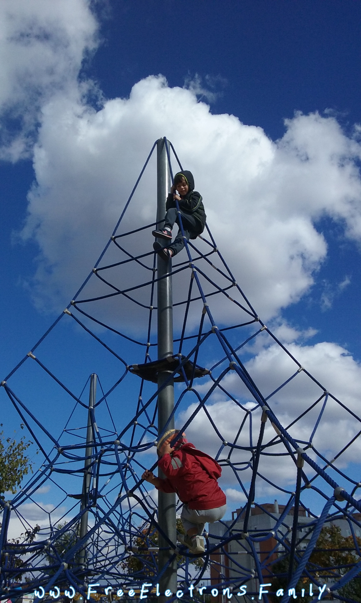Two young children at free play, climbing a tower connected with web-like ropes, at "Ciudad de los Ninos"  (City of Children).  Background Andalucian blue sky with some cumulus clouds.