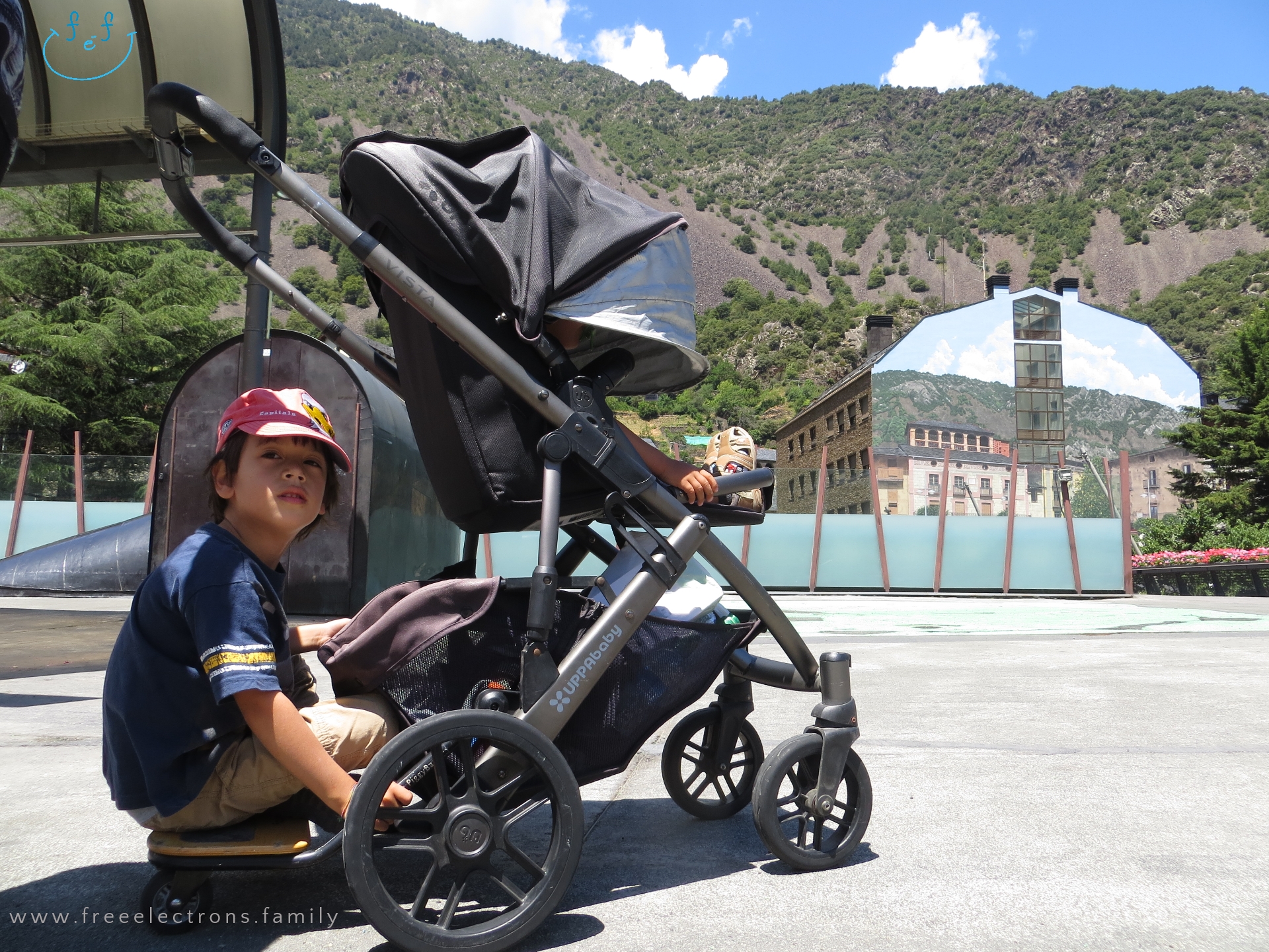 #FreeElectrons.Family - camping road trip Europe, Andorra by Uppababy stroller/pram.

Two young boys on a stroller in a wide open square, with a painted building that blends into a mountain background.

Text reads: www.freeelectrons.family