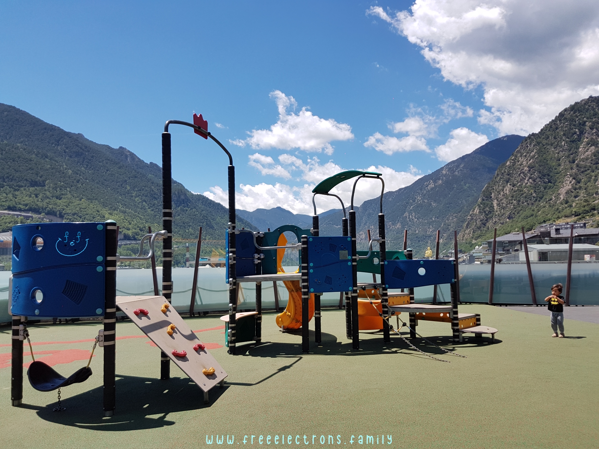 #FreeElectrons.Family - camping road trip Europe, Andorra centro playground.

A lone young boy in a modern playground with the mountains and blue bright sky in the background.

Text reads: www.freeelectrons.family