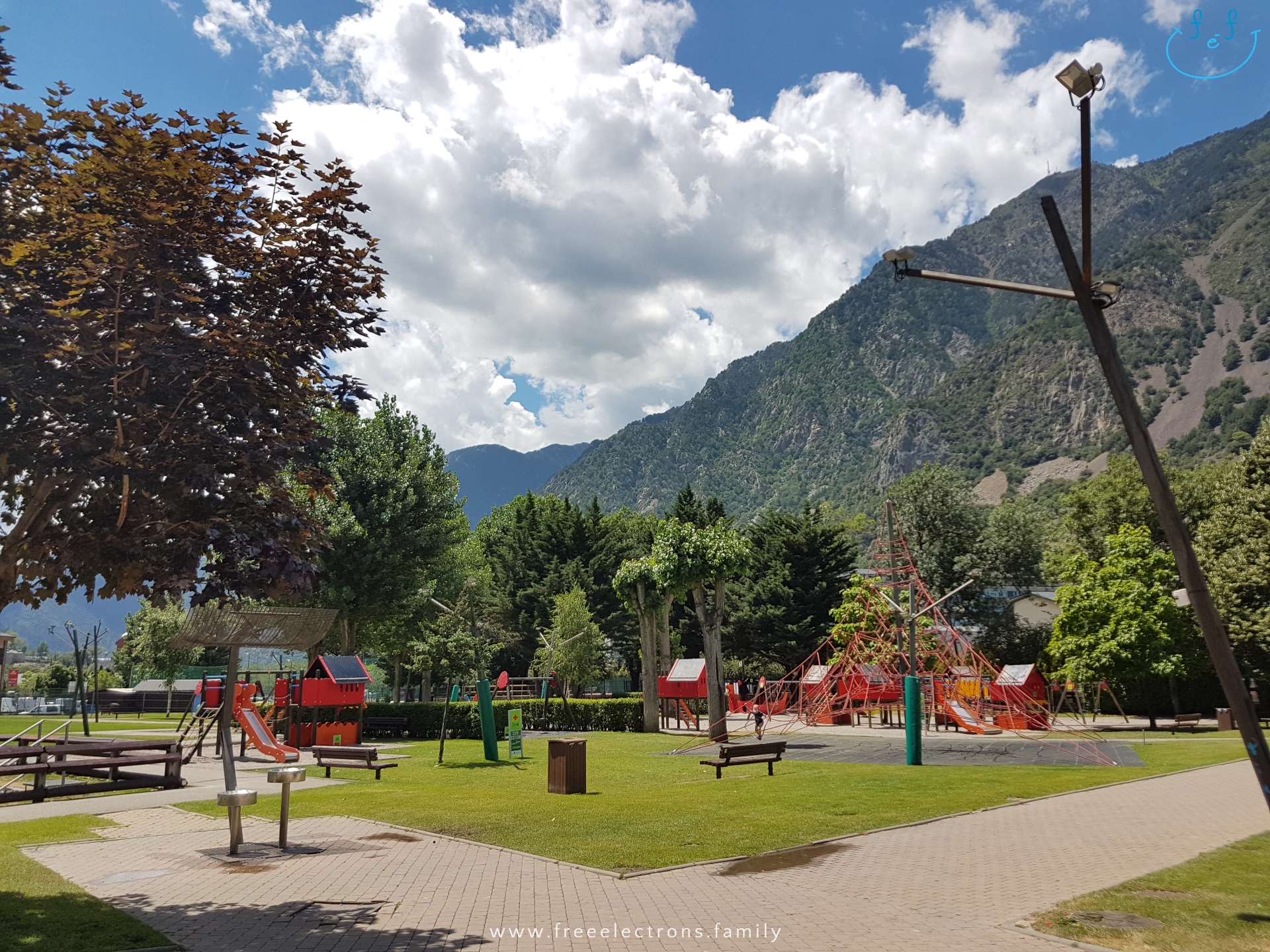 #FreeElectrons.Family - camping road trip Europe, Andorra playground.

A clean, modern and colorful playground in a park with lots of trees and mowed grass, with the mountainside and cloudy bright blue skies in the background.

Text reads: www.freeelectrons.family