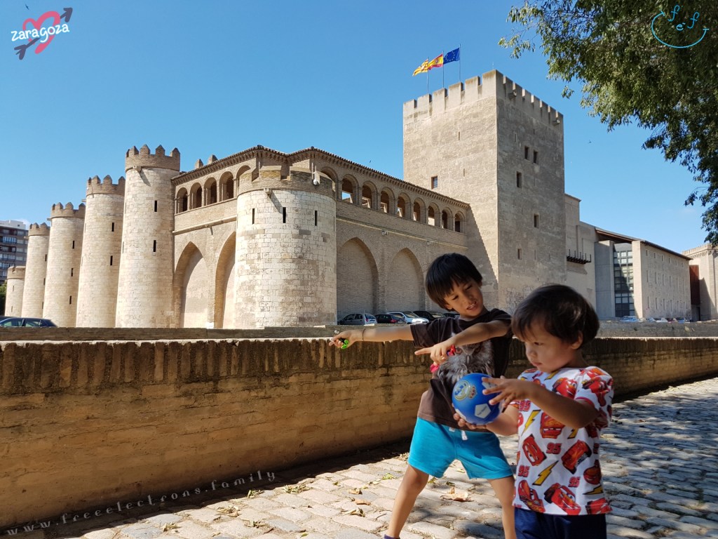 Two young boys at free play at the tree-lined park just outside the Aljaferia. The EU, Spanish and Aragon flags fly above the castle-fortress in the background. Text reads: Zaragoza with a symbol of a heart and an arrow behind it, and "www.freeelectrons.family".
