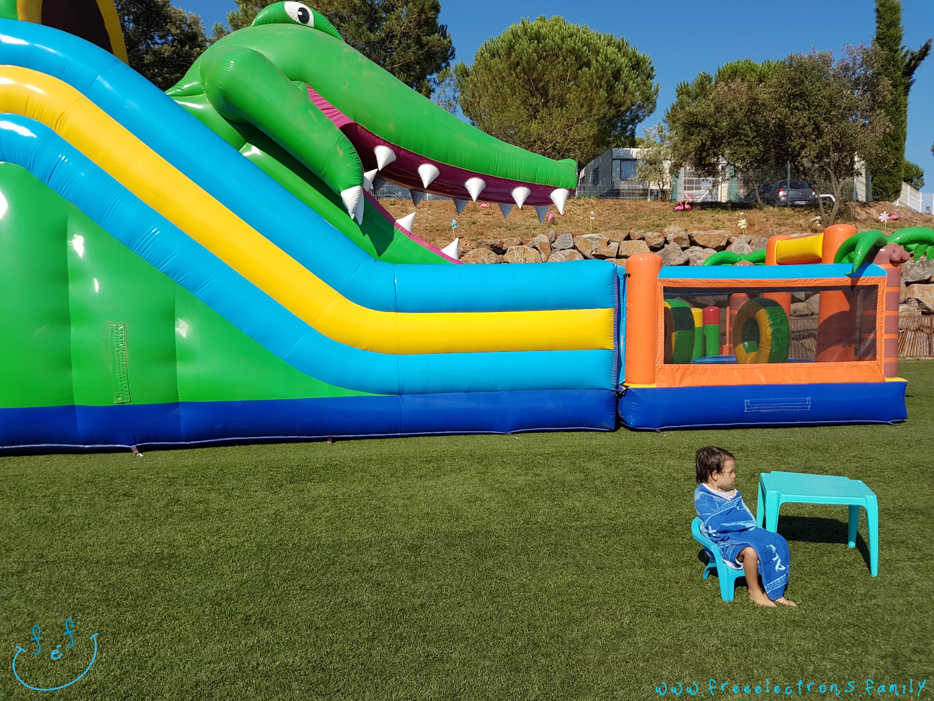 A young boy wrapped in a towel, sitting on a kids chair on green grass, next to a huge crocodile-themed, jump-and-slide,  inflatable play structure.

#FreeElectrons.Family - camping road trip Europe, Agde France.