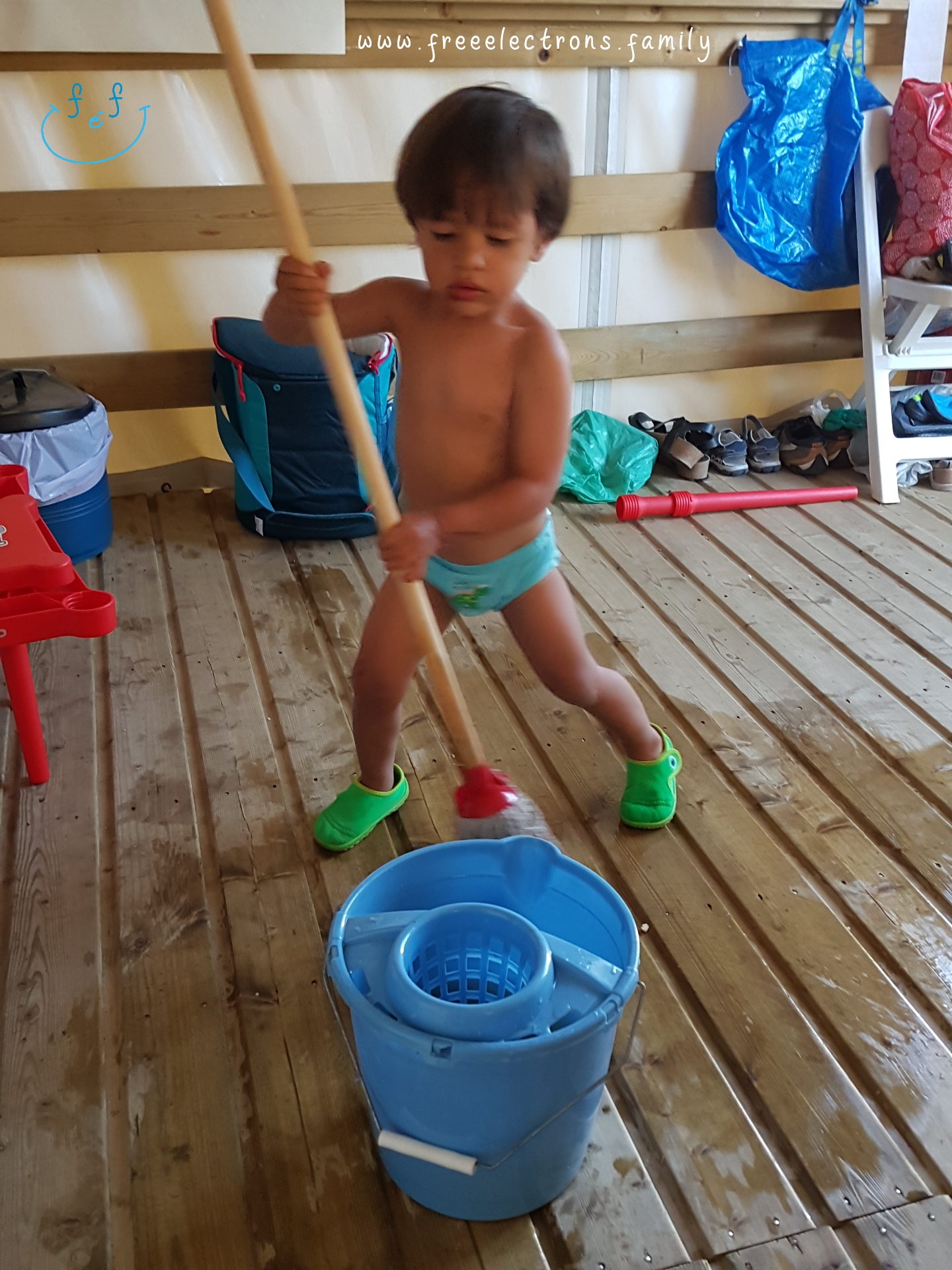 A young boy, shirtless, cleans up with a broom in a camping cabin.

#FreeElectrons.Family - camping road trip Europe, Agde France.