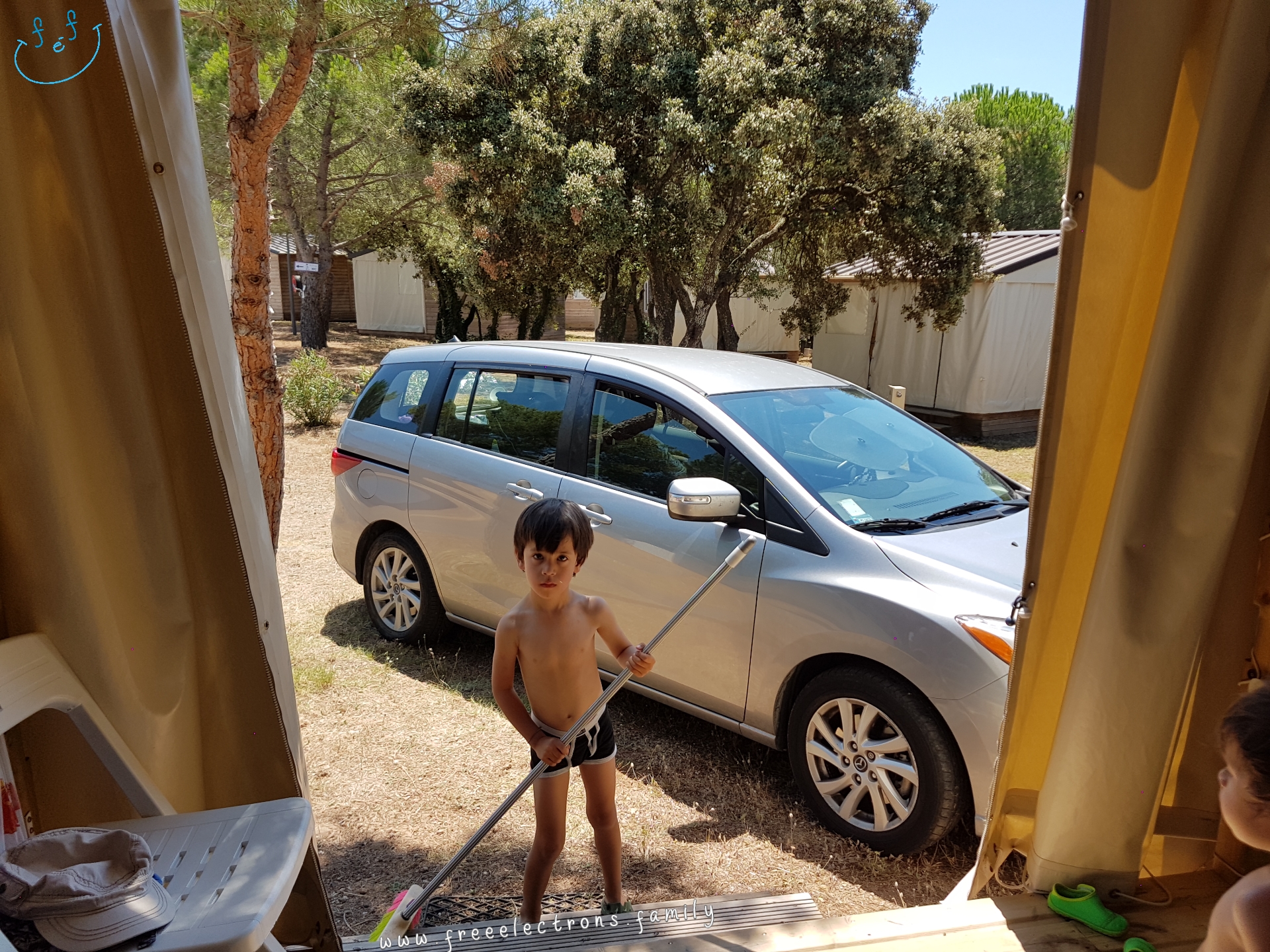 A young boy, shirtless, cleaning up with a broom in front of  camping cabin.  A younger boy looks at him from a patio.

#FreeElectrons.Family - camping road trip Europe, Agde France.