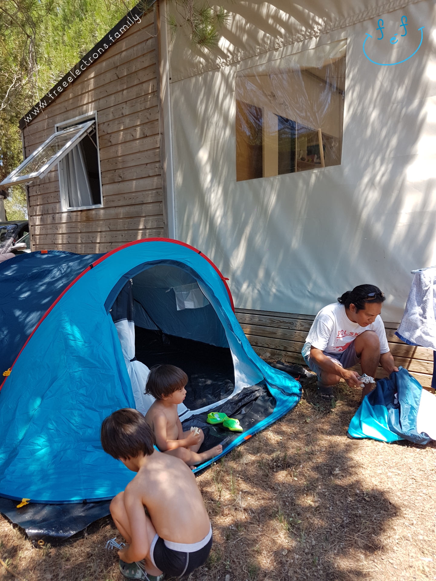 A father with 2 young kids setting up tent a tent next to a cabin.

#FreeElectrons.Family - camping road trip Europe, Agde, France.