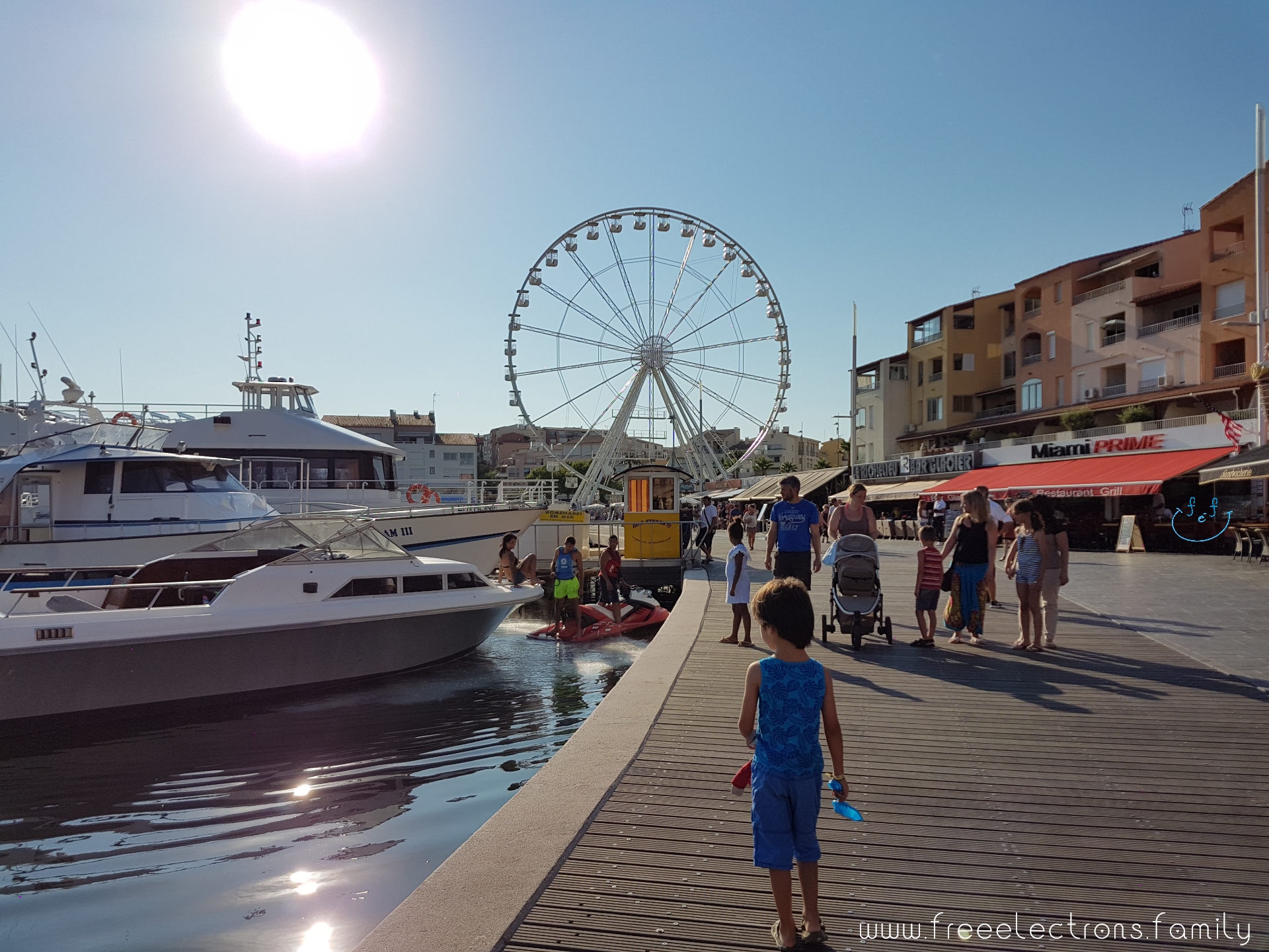 A young boy walks on a boardwalk of a marina, with others in the background, ahead of him.  There are modern boats on the water and a grand ferris wheel up ahead.

#FreeElectrons.Family - camping road trip Europe, Cap d'Agde France.