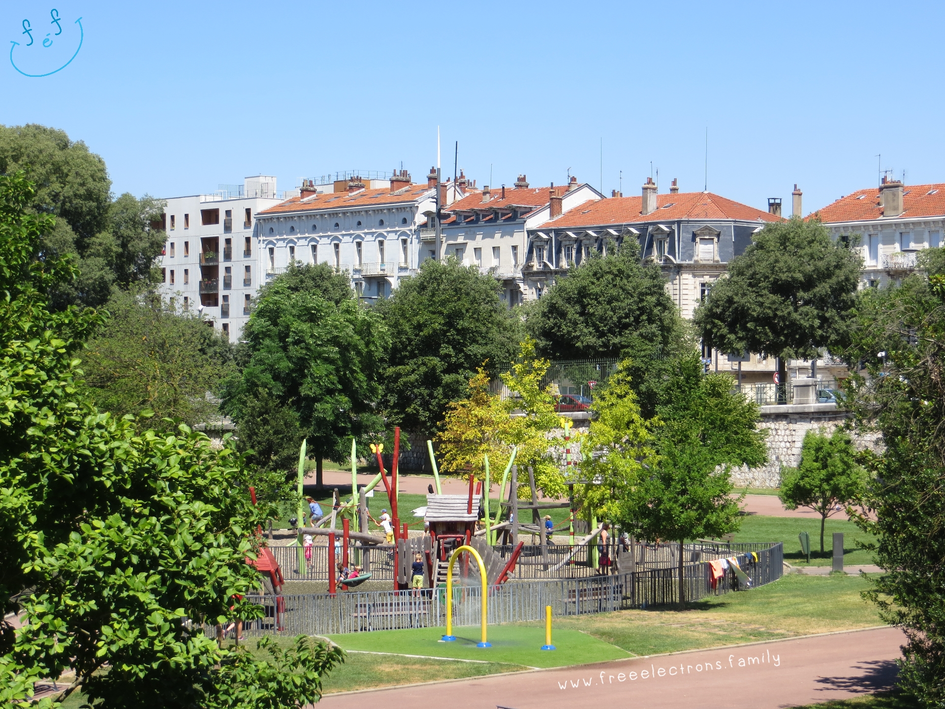 One of the playgrounds at Parc Jouvet, surrounded by trees, with the city's buildings in the background.

#FreeElectrons.Family - camping road trip Europe, Valence.