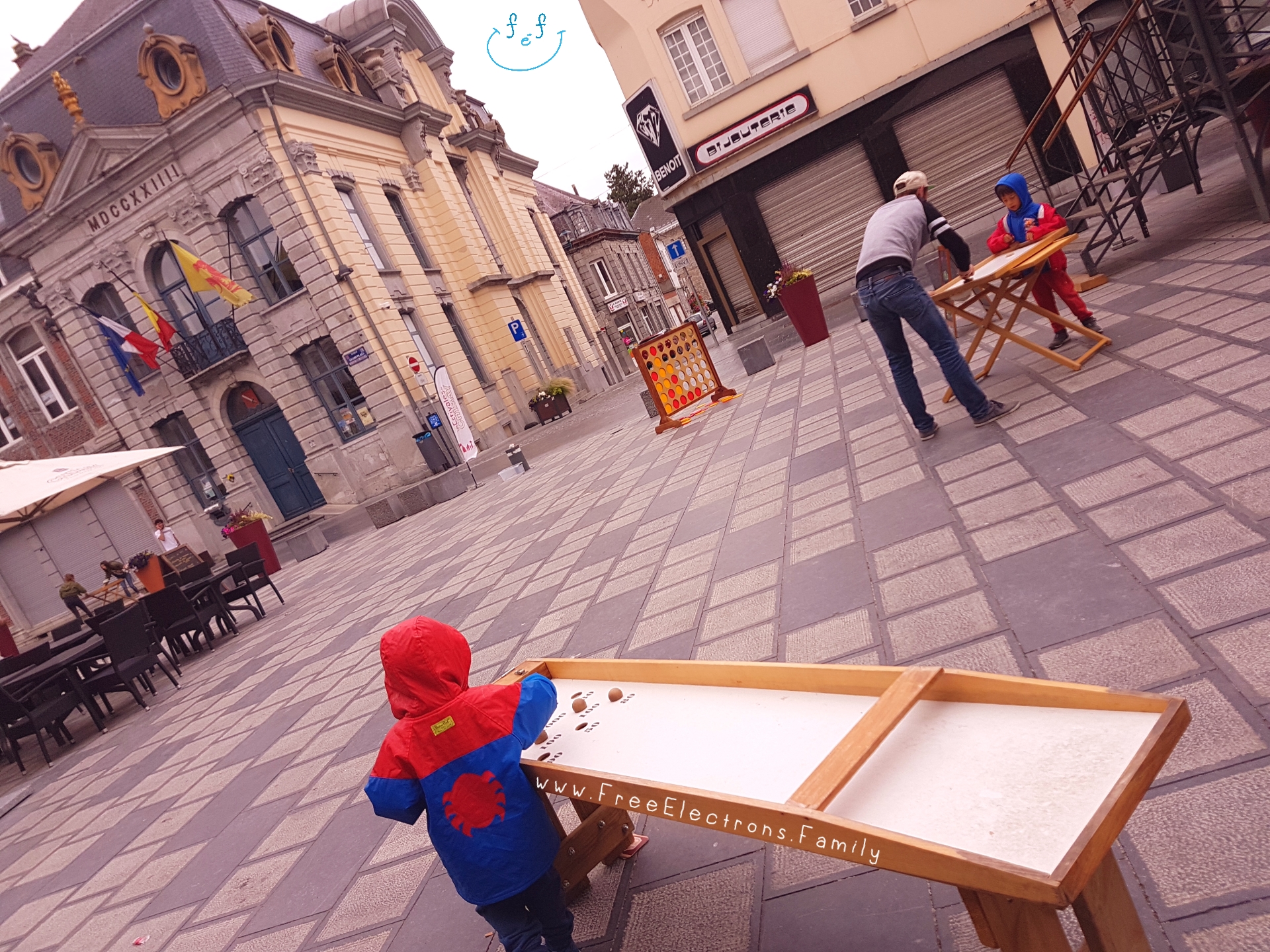 Other old-school games at Chimay town square.  Two boys in raincoats; one plays with an adult.

www.FreeElectrons.Family - camping road trip Europe 2.