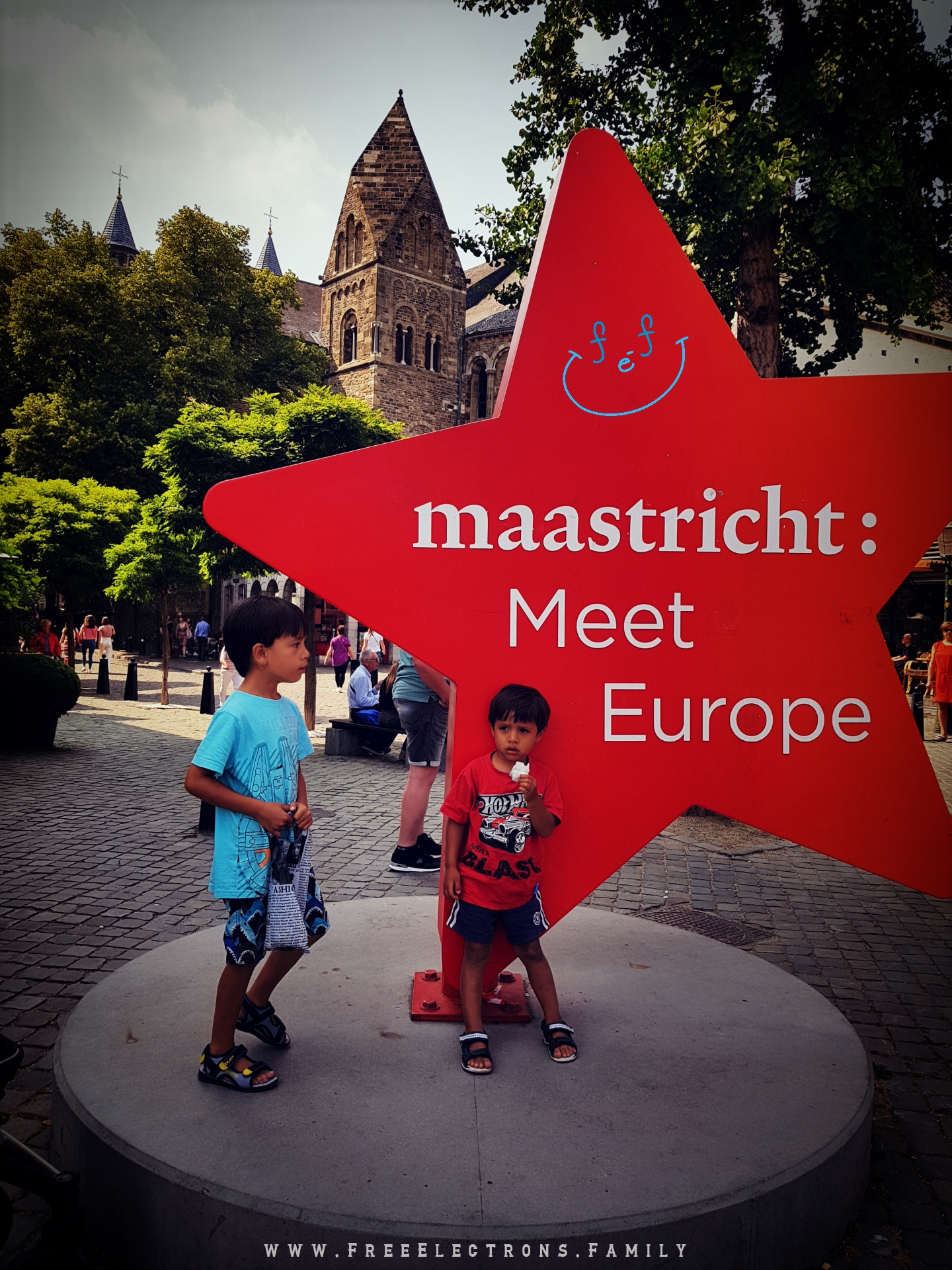 Two young kids, looking tired, at a sign that reads "maastricht: Meet Europe.

#FreeElectrons.Family - camping road trip Europe Maastricht