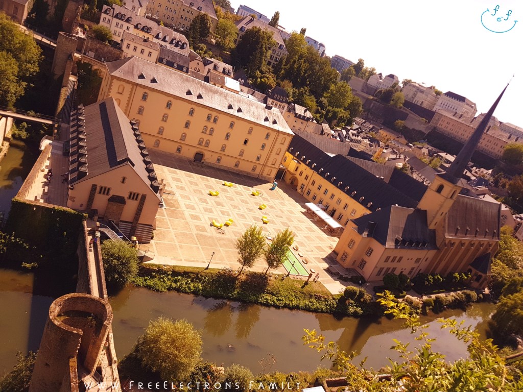 View from the rocky Casemates du Bock overlooking a valley with a courtyard of an abbey, the Alzette river and an old bridge.

Text on picture reads: www.FreeElectrons.Family with an inserted smiley face (custom icon) at a top corner. 