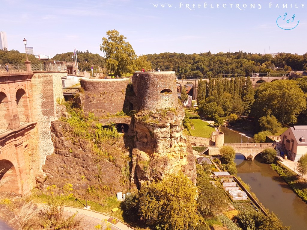 View of the rocky Casemates du Bock over a valley and a river with an old bridge.

Text on picture reads: www.FreeElectrons.Family with an inserted smiley face (custom icon) at the top corner. 