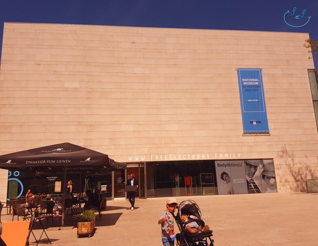Two young boys (one standing, the other sleeping in a pram) in front of a windowless modern building.  An outdoor cafe at the entrance.

Text on picture reads: www.FreeElectrons.Family with an inserted smiley face (custom icon) at a top corner. 