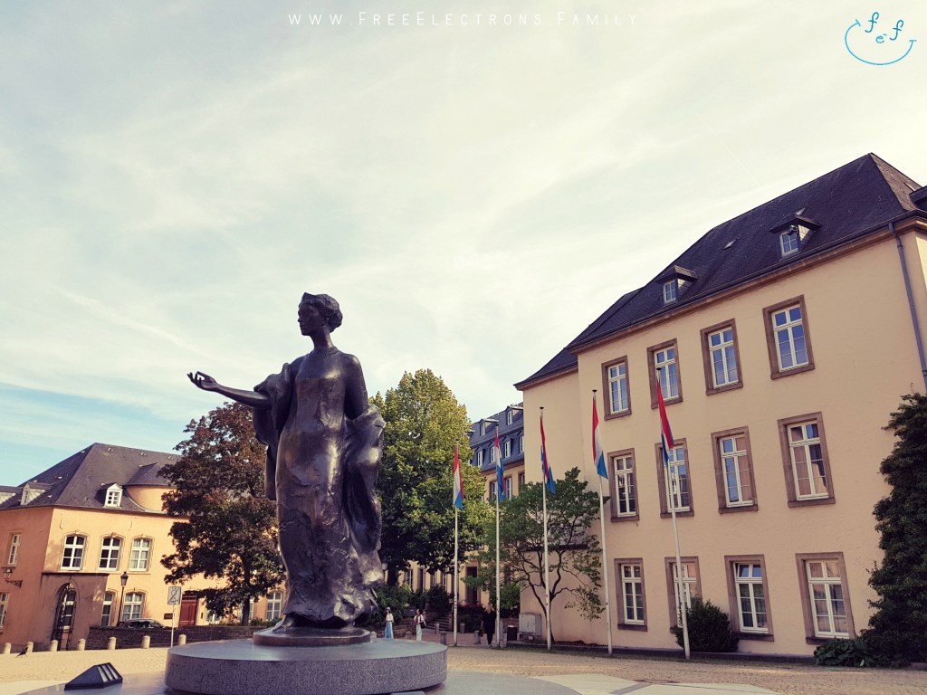 A front-facing bronze statue of the Grand Duchess Charlotte--the beloved monarch of Luxembourg-- in an empty square/plaza, under blue-grayish sky with an inserted smiley face (custom icon) at the bottom corner. 

In the background are buildings; one of which is a 3-storey building with the country's blue-white-red flags in front of it.

Text on picture reads: www.FreeElectrons.Family
