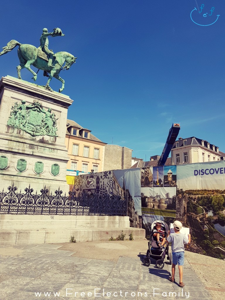 Two young boys at the foot of a greenish-colored copper-based statue of a man on a horse, under blue sky with an inserted smiley face custom icon at the top corner.

One of the boys is looking at a map, while the other sits in a pram/stroller.

Text on picture reads: www.FreeElectrons.Family
