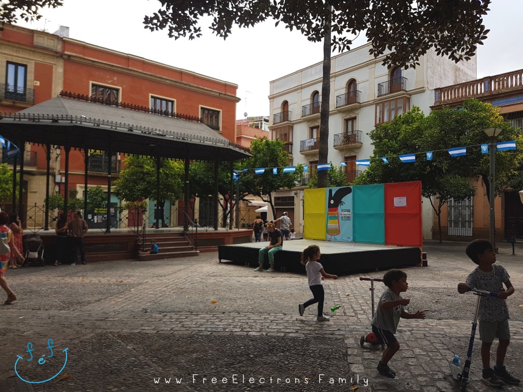 Children playing while waiting for an outdoor puppet show in a Spanish plaza, under cloudy skies.

Caption reads:  Waiting for the puppet show at Plaza del Banco, Jerez de la Frontera, International Puppet Festival (Festival Internacional de Títeres) in Septembers.

www.FreeElectrons.Family - what to do see in Jerez in September.