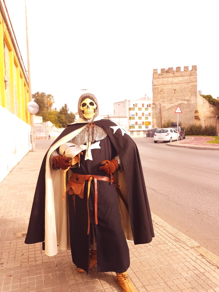 A skeleton of a knight in full armour, carrying a a helmet and a sword, posing in a modern (paved) street with part of an old medieval wall-tower fortification in the background, in broad daylight.