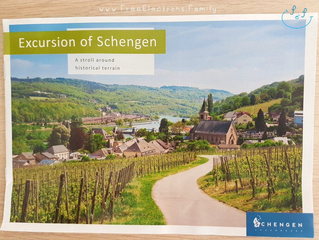 A photo of a brochure titled "Excursion of Schengen: a stroll around historical terrain" with a picturesque view of small village overlooking the river and the vineyards on the other side.

Other text on picture reads: "Schengen" and www.FreeElectrons.Family.