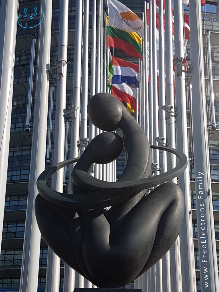 "Europe a Coeur" monument, 2 abstract people hugging--symbol of the European Union, at the entrance of a steel-and-glass building with flags in the background.

Text on photo reads www.FreeElectrons.Family