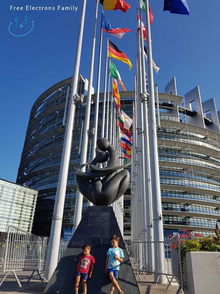 Two young kids stand at a "Europe a Coeur"monument at the entrance of a steel-and-glass building with flags in the background.

Text on photo reads www.FreeElectrons.Family