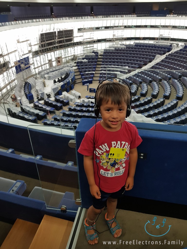 A young boy stands in the plenary hall (in the auditorium) of the European Parliament, with the circular members's section in the background.

Text on photo reads www.FreeElectrons.Family