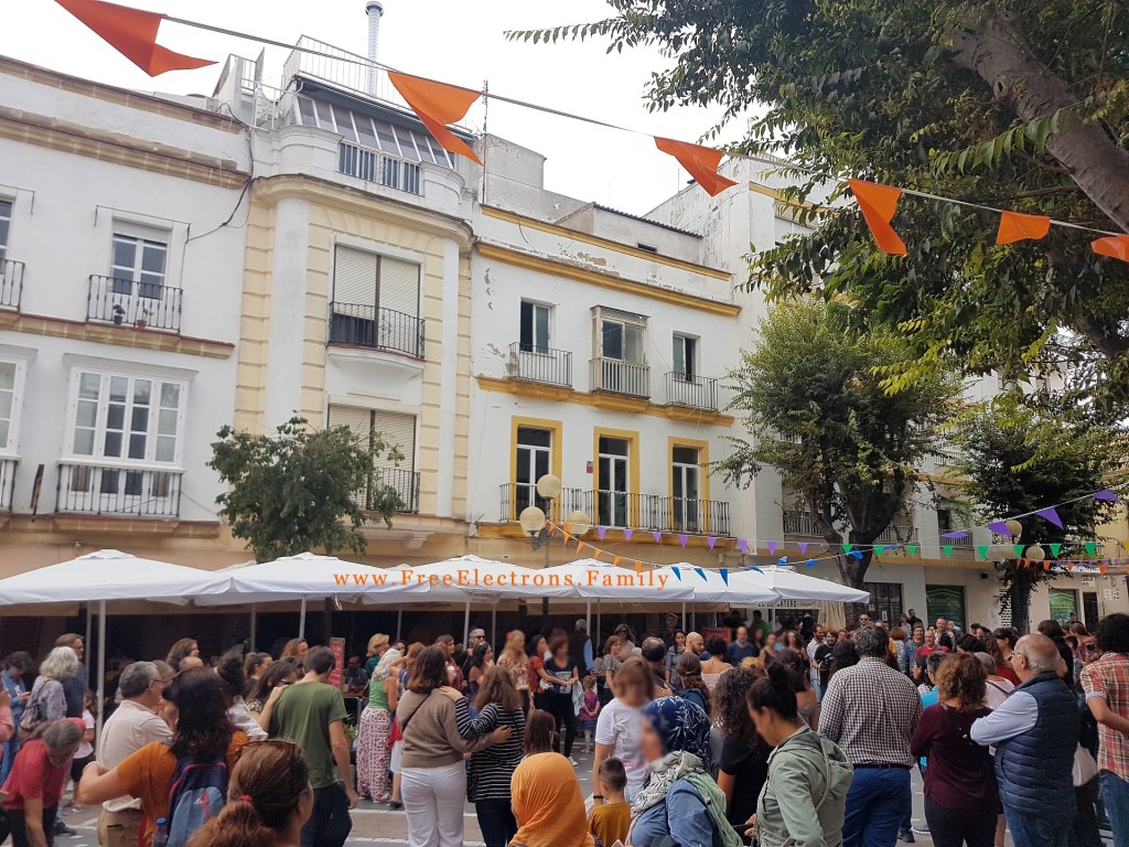 A Spanish plaza/square filled with dancing people and spectators, with 3-storey buildings with balcony and some trees in the background.

Text on photo reads: www.FreeElectrons.Family