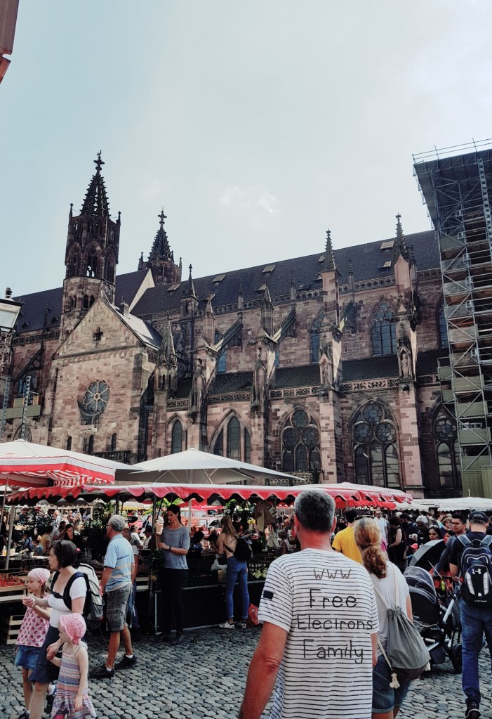 A busy open market with the Freiburg cathedral in the backdrop.

A shirt in the foreground reads: 
 www.FreeElectrons.Family