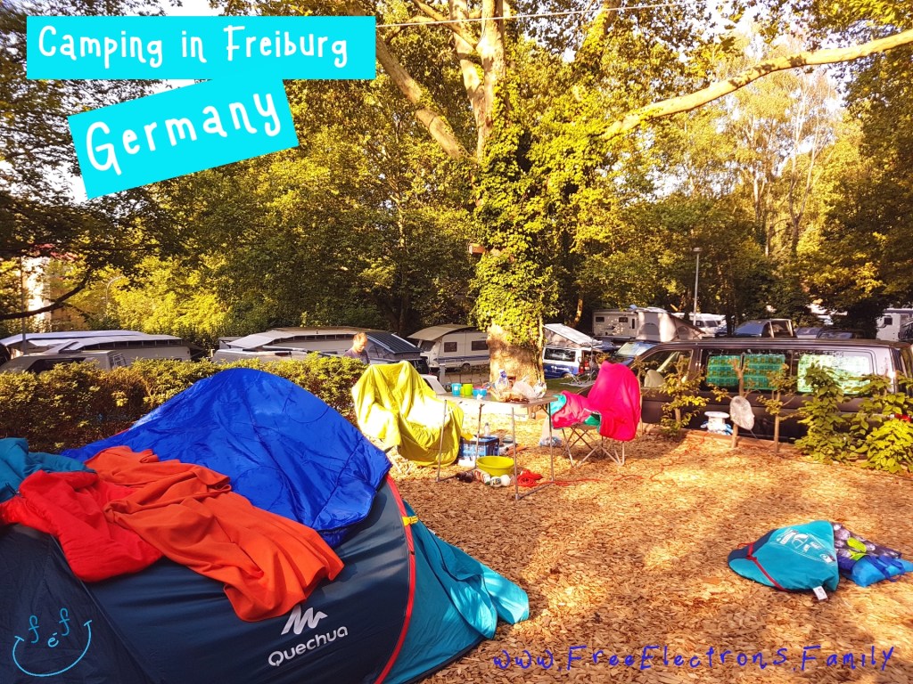 Camping pitch with a tent, tables, chairs and other stuff; camping vans and shadeful trees in the background.

Text reads: 
Camping in Freiburg, Germany
www.FreeElectrons.Family