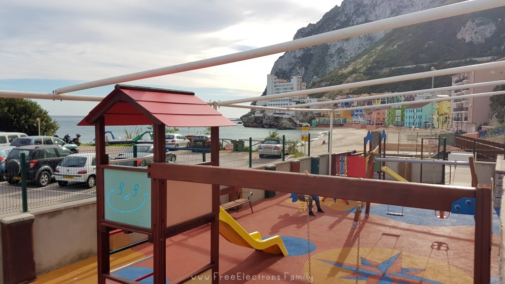 A clean and colourful playground on Caleta Bay beach with colorful houses at the foot of the rock mountain of Gibraltar in the background.

Text reads: www.FreeElectrons.Family