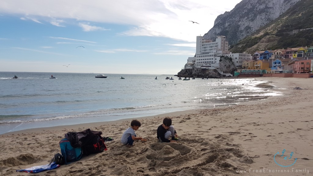 Two young boys playing at the beach on Caleta with colorful houses at the foot of the rock mountain of Gibraltar in the background.

Text reads: www.FreeElectrons.Family