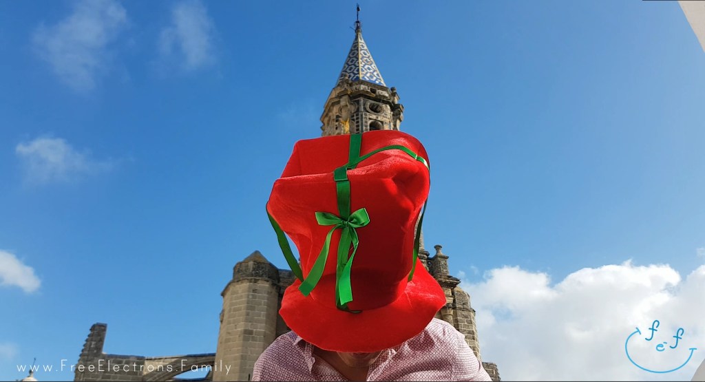 A father with a huge Christmas hat covering his face with an old church and its bell tower behind him, under Andalusian blue sky.

www.FreeElectrons.Family