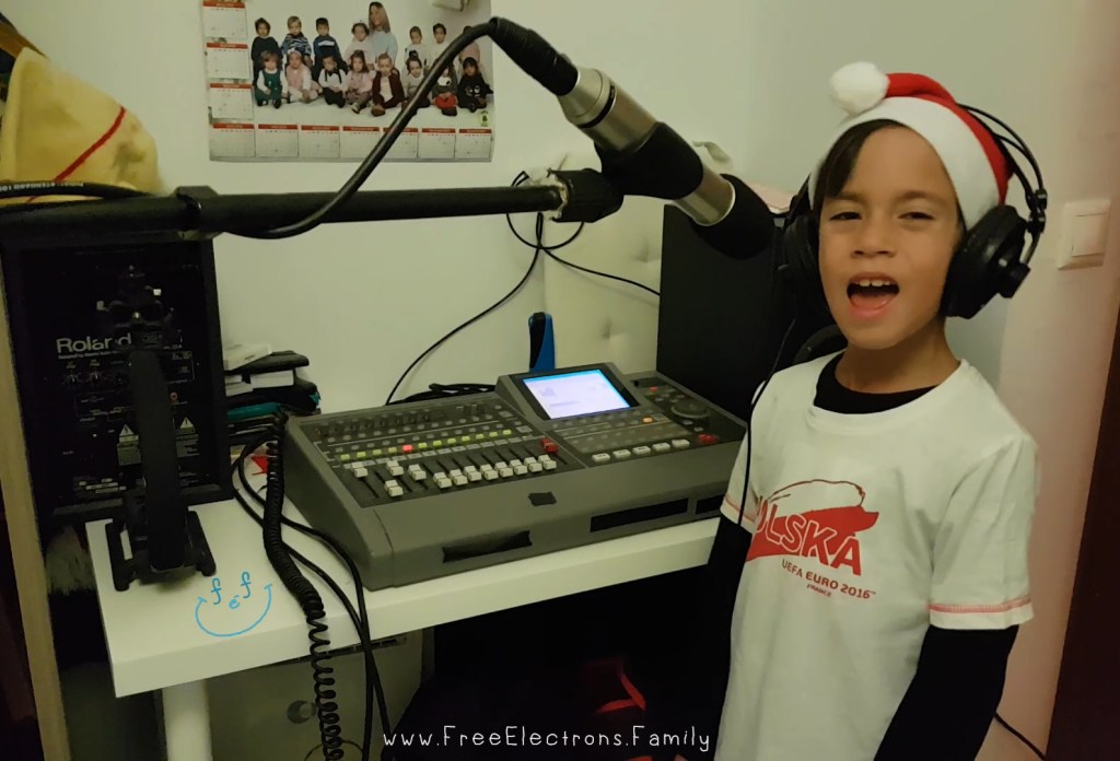A young boy  in a white "Polska" shirt with a Santa Claus hat and headphones on singing in front of a microphone in a home recording studio.

www.FreeElectrons.Family
