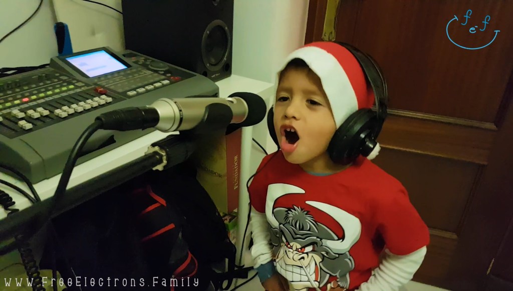 A young boy in a red shirt with a Santa Claus hat and headphones on singing in front of a microphone in a home recording studio.

www.FreeElectrons.Family