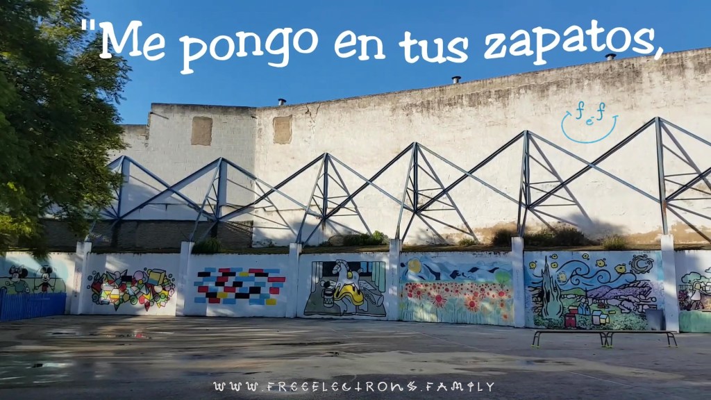 An outdoor patio with various paintings on the walls.  Text reads in Spanish, "Me pongo en tus zapatos" (I put myself in your shoes).  
www.FreeElectrons.Family