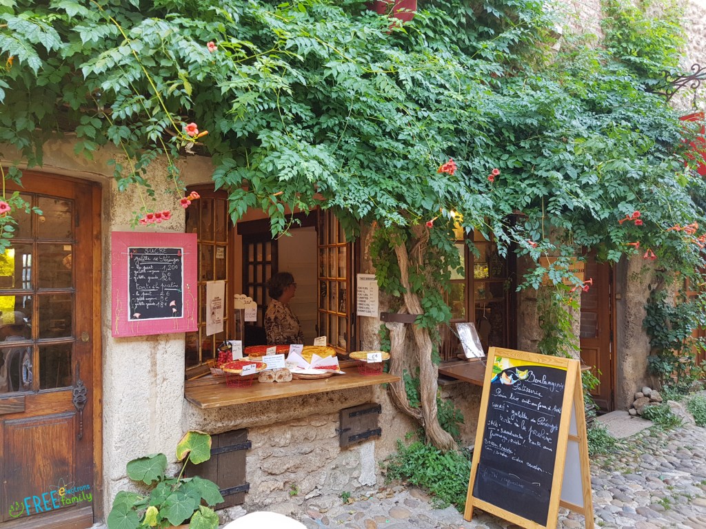 A small shop with fully grown vines overhanging windows open and offering pizzas and other foodstuff with blackboards listing names and prices.