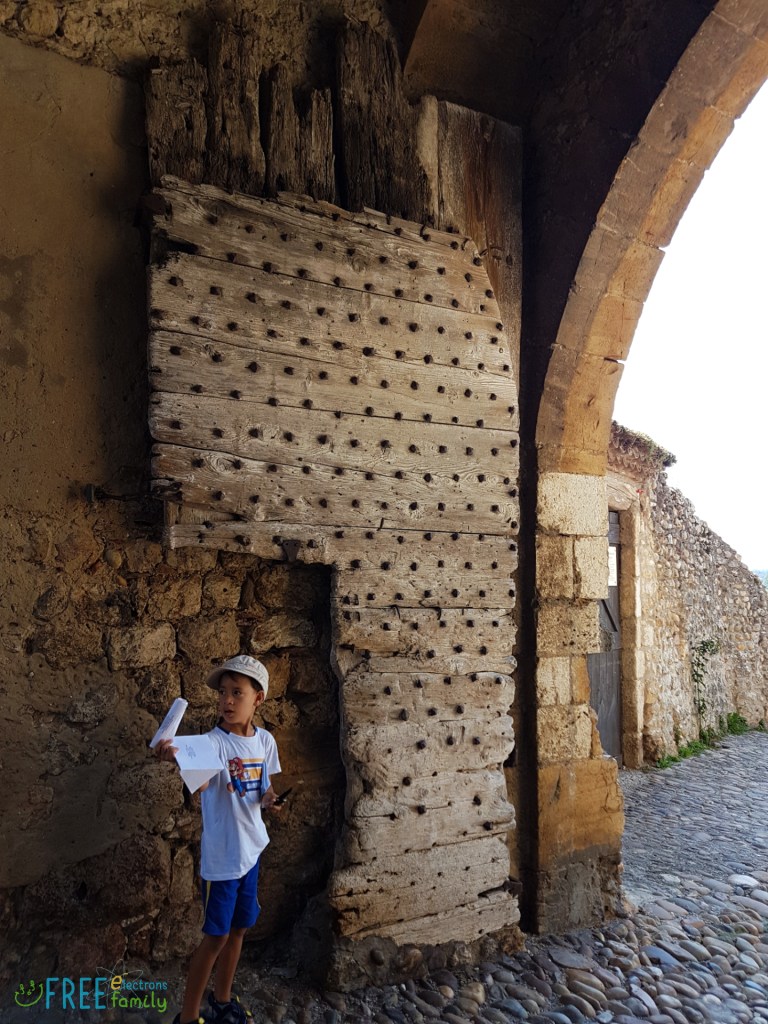 A young boy, looking away, holds a white paper by the broken wooden gate, under a stone archway entrance of a cobblestone village.

www.FreeElectrons.Family