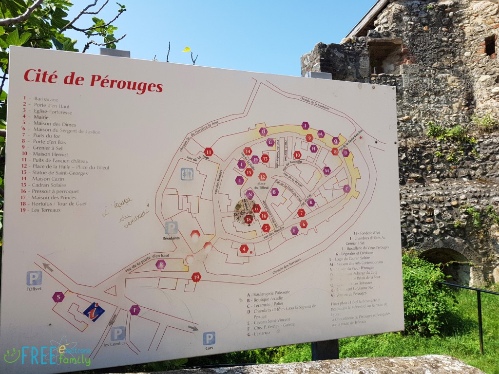A map of the Cite de Perouges with part of a stone house in the background, grass and blue sky.