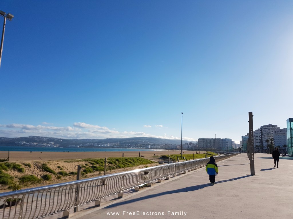 A lone boy walks away along a wide promenade with beach and mountain in the background.