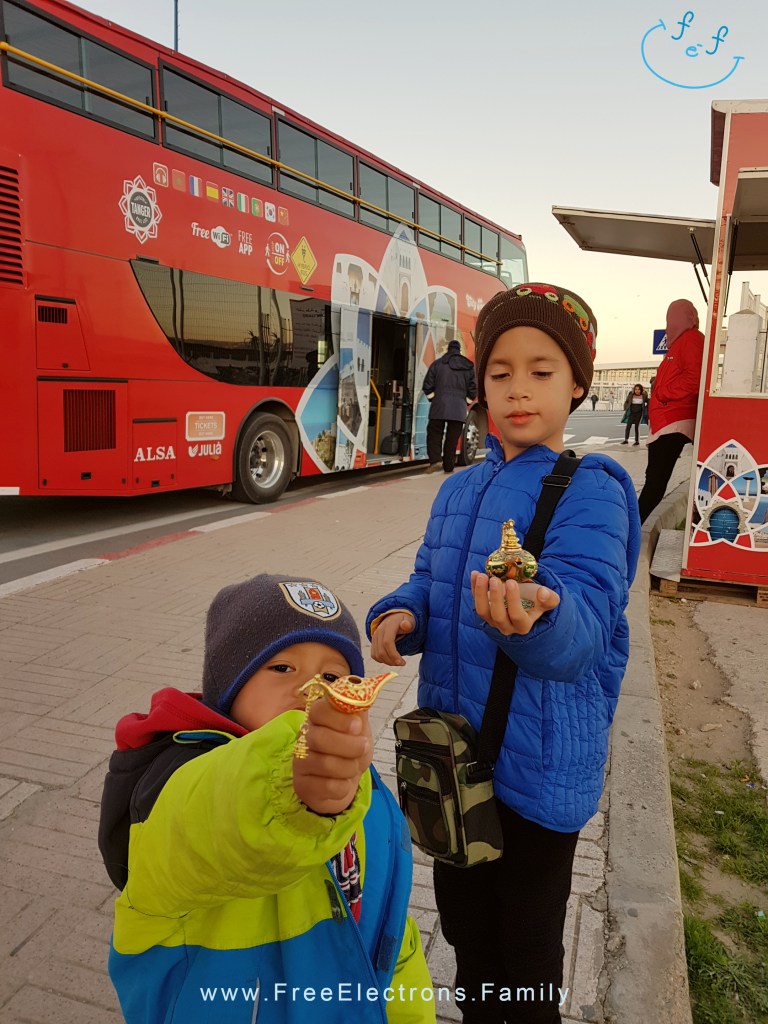 Two young boys in winter clothes show off their little souvenirs in front of a red double-decker bus.