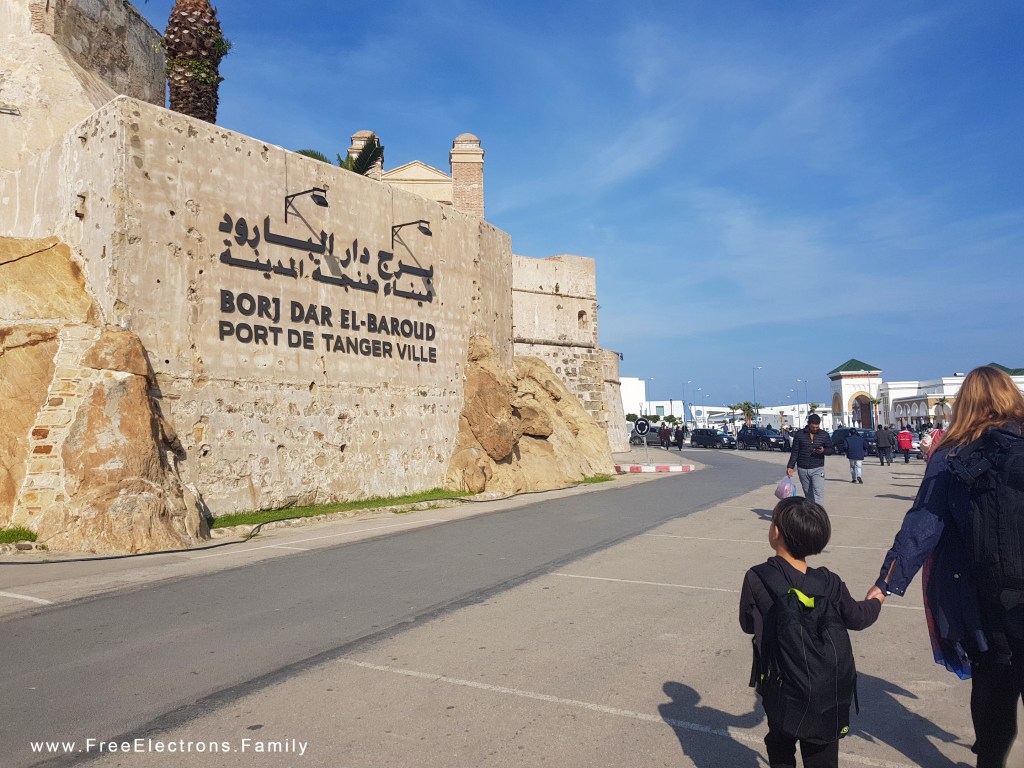 A young boy, holding his mother's hand, looks at an ancient wall with modern writing in Arabic, French and Berber that reads "Port de Tanger Ville". 
