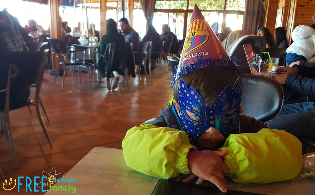 A young boy with a carnival mask and a FC Barcelona hat sits in a restaurant.